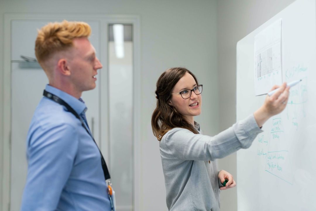 zwei Personen stehen vor einem beschriebenen Whiteboard. Die Frau zeigt auf das Board mit einem Stift und scheint etwas zu erklären.