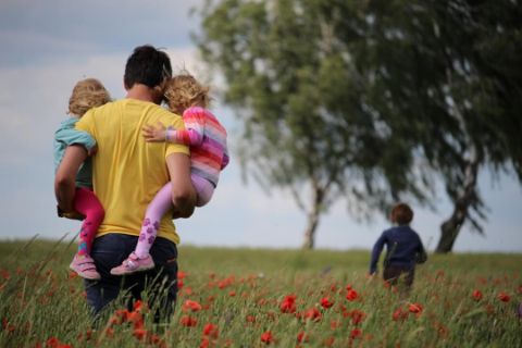 Vater mit zwei Kindern auf dem Arm von hinten auf einer Wiese mit Mohnblumen und einem weiteren Kind weiter vorne