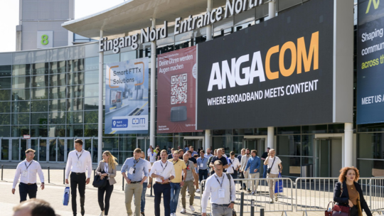 Das Bild zeigt den Eingang Nord einer Messehalle mit einem großen Schild, das "ANGA COM" und den Slogan "WHERE BROADBAND MEETS CONTENT" anzeigt. Eine Gruppe von Messebesuchern bewegt sich in Richtung Eingang. Im Hintergrund sind weitere Schilder und Werbebanner zu sehen, die auf verschiedene Aussteller und Themen hinweisen. Die Veranstaltung ANGA COM 2024 konzentriert sich auf Breitband- und Mediendistribution. Das Wetter ist sonnig, und die Besucher tragen überwiegend legere Geschäftskleidung.