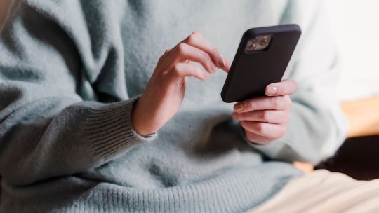 Eine Person in einem hellgrünen Pullover und beigefarbenen Hosen sitzt und hält ein Smartphone in beiden Händen. Der Fokus liegt auf den Händen der Person, die das Telefon bedienen. Das Gesicht der Person ist nicht im Bild zu sehen, da der Ausschnitt nur den Oberkörper und die Hände zeigt. Die Person scheint konzentriert auf das Gerät zu schauen und es zu benutzen.