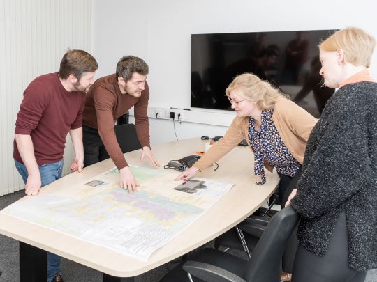 Vier Personen stehen um einen Tisch mit einem Netzplan und zeigen darauf