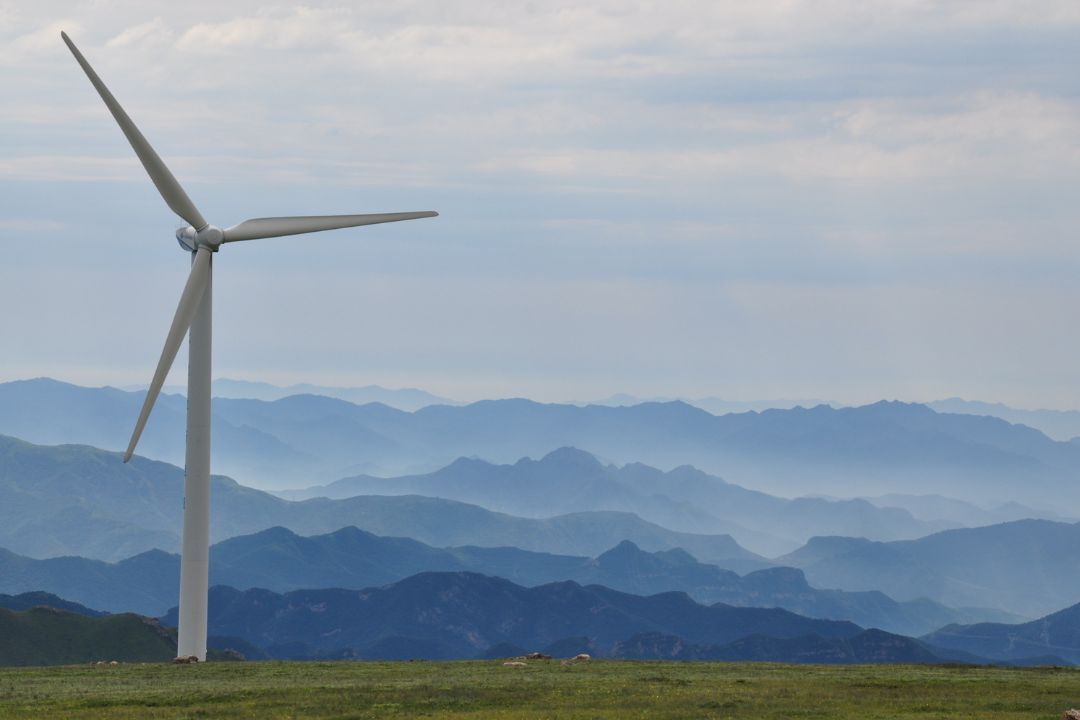 Das Bild zeigt eine einzelne Windkraftanlage, die auf einem grünen Hügel steht. Im Hintergrund erstreckt sich eine beeindruckende Berglandschaft mit mehreren Bergketten, die in verschiedenen Blautönen verblassen und von einem bewölkten Himmel überragt werden. Die Szene vermittelt ein Gefühl von Ruhe und Weite inmitten der Natur.