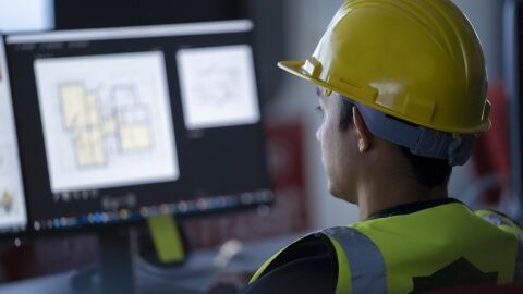 Mann mit Bauhelm, der auf zwei Monitore blickt und seine Aufgaben in der Bauüberwachung wahrnimmt