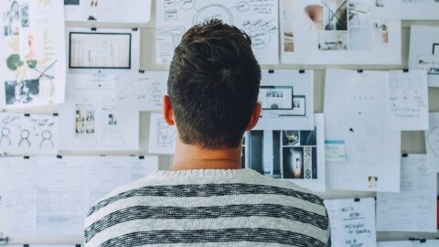 Mann von hinten, der vor einer Pinnwand mit einer Vielzahl von Blättern und Informationen steht und im Ausschreibungs- und Vergabemanagement den Überblick verloren hat