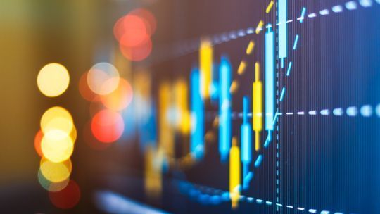 Das Bild zeigt eine unscharfe Darstellung eines Diagramms mit Kerzencharts auf einem Bildschirm. Die Kerzen sind in Blau und Gelb dargestellt. Im Vordergrund sind unscharfe, bunte Lichter zu sehen. Das Bild hat eine verschwommene Ästhetik.