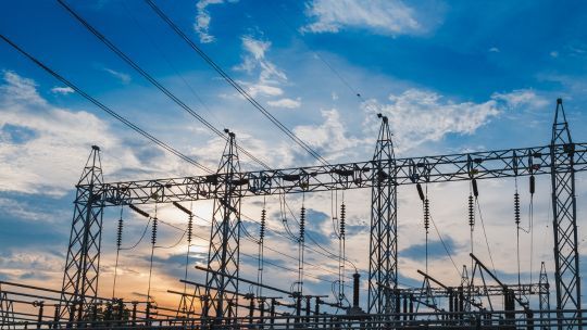 Eine elektrische Umspannstation mit mehreren Stahlkonstruktionen und Hochspannungsleitungen, die sich vor einem bewölkten Himmel bei Sonnenuntergang oder -aufgang befinden. Die Kabel und Isolatoren sind deutlich sichtbar, und die Szene vermittelt die Bedeutung der Energieinfrastruktur. Die Farben des Himmels reichen von Blau bis hin zu warmen Gelb- und Orangetönen.