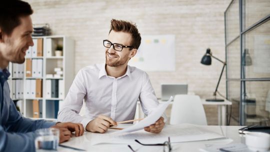 Zwei lächelnde Geschäftsmänner in einem modernen Büro sitzen an einem Tisch und diskutieren Dokumente. Einer der Männer hält Papiere in der Hand und erklärt etwas, während der andere aufmerksam zuhört. Im Hintergrund sind Regale mit Ordnern und eine Schreibtischlampe zu sehen.