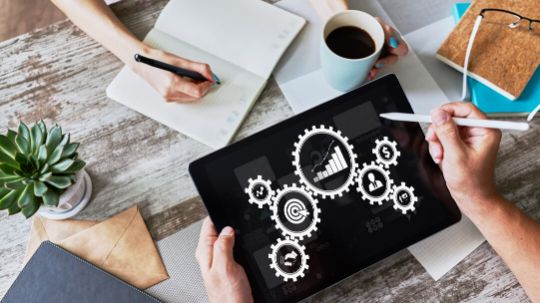 Arbeitstisch von oben, an dem aus zwei Richtungen jeweils zwei Hände sowie eine Pflanze, eine Tasse Kaffee und ein Tablet zu sehen sind. Das Tablet zeigt eine Grafik zur Prozessoptimierung mit Zahnrädern und Daten.