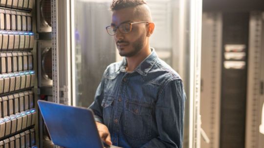 Administrator im Serverraum, der auf einem Laptop die IT-Infrastruktur und die IT-Sicherheit überwacht