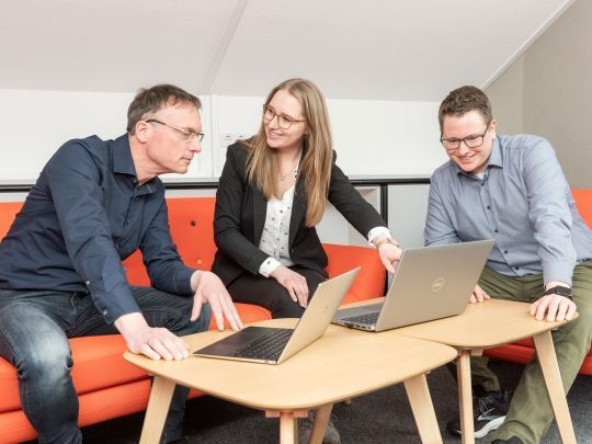 Drei Personen stehen um einen Tisch mit zwei Laptops und reden zusammen