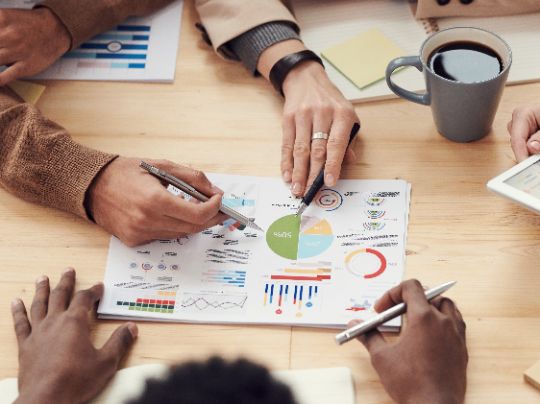Das Bild zeigt Dokumente mit statistischen Analysen, auf die Hände verschiedener Menschen deuten. Es steht symbolisch für die Markterkundung im Rahmen der Graue-Flecken-Förderung.