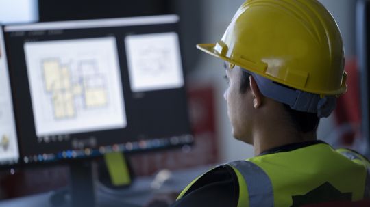 Mann mit Bauhelm auf dem Kopf sitzt vor einem Computerbildschirm mit Netzplan