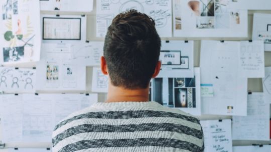 Mann von hinten, der vor einer Pinnwand mit einer Vielzahl von Blättern und Informationen steht und im Ausschreibungs- und Vergabemanagement den Überblick verloren hat